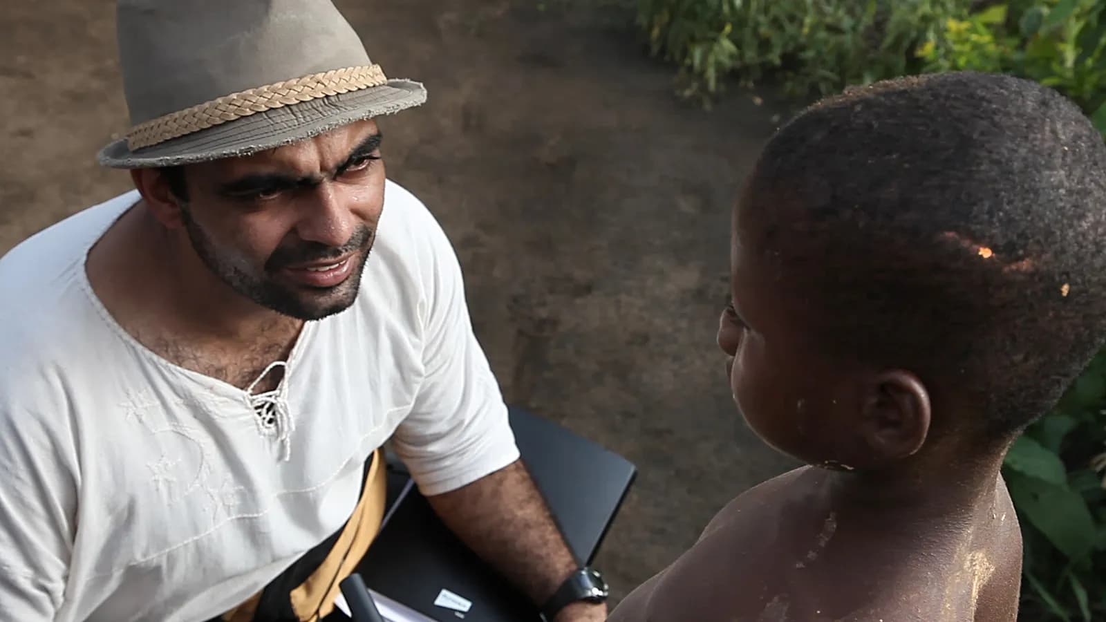 A man in a fedora hat and white shirt is talking to a young child with their back to the camera.