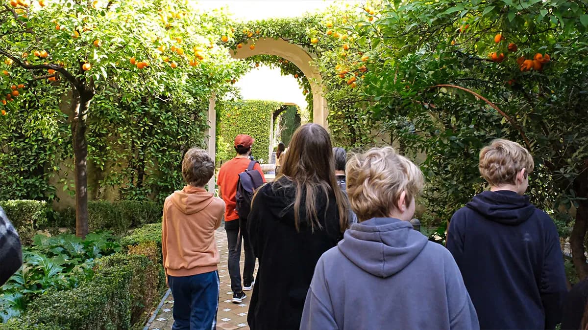 A group of young people walking through a sunny garden with lush green foliage and orange trees laden with fruit, towards an arched walkway.